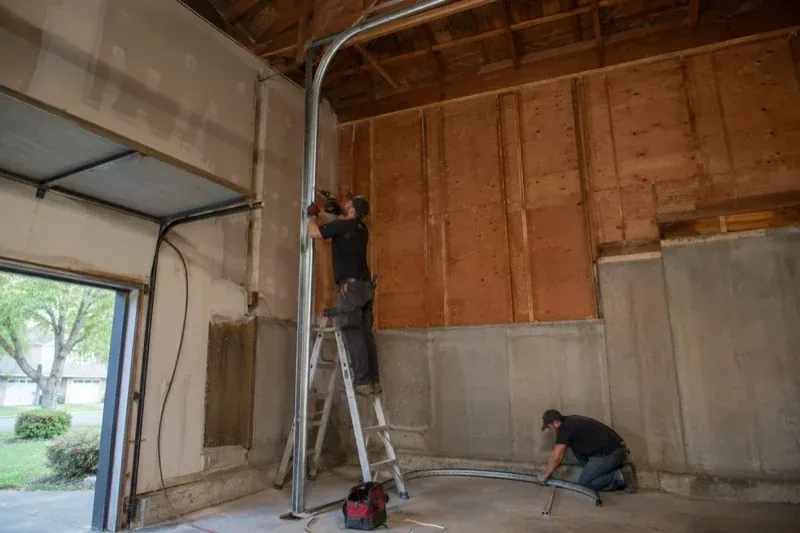 One person on a stepladder working beside a tall curved garage-door track while another kneels beside loose track sections in an unfinished garage. — High-Lift Garage Door Conversion, Toronto